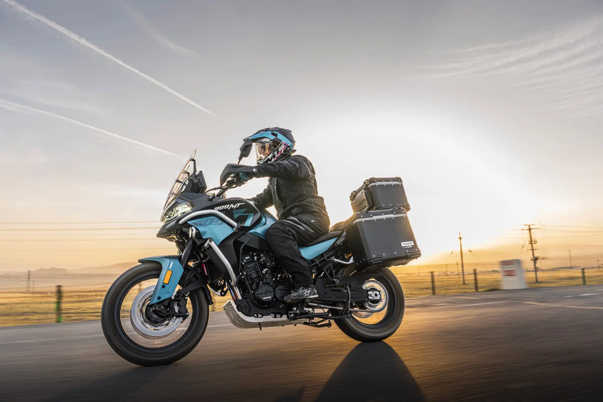 A person wearing protective gear rides a light blue 800MT-X adventure motorbike with panniers on a tarmacked road at sunrise or sunset.