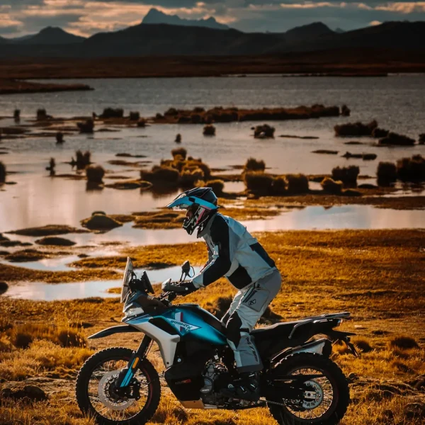 A person wearing motorcycle gear rides an 800MT-X adventure motorcycle across a grassy landscape near a body of water at sunset.