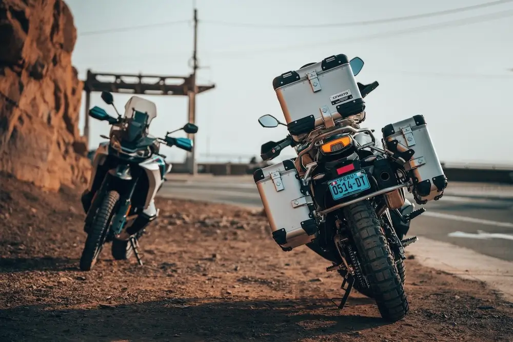 Two adventure motorcycles with panniers equipped with CFMOTO T-Box are parked on a dirt roadside next to a rocky cliff, with a torii gate and power lines visible in the background.
