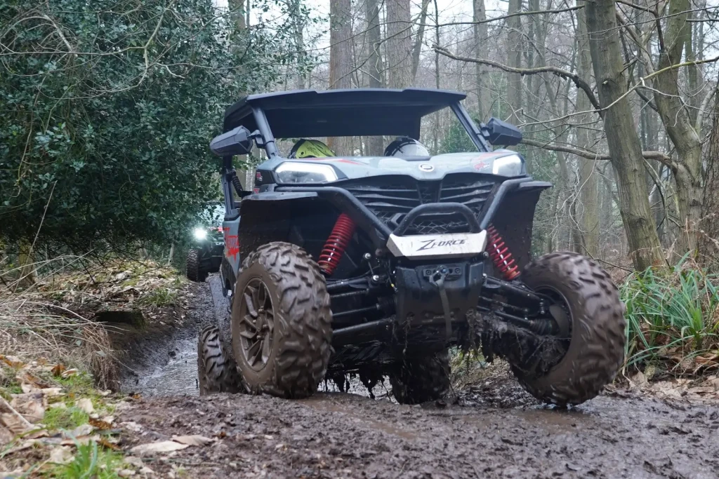 A rugged off-road vehicle navigates a muddy forest trail with ease, its noticeable suspension and large tires reminiscent of a farm quad, cutting through the dense trees and lush foliage.