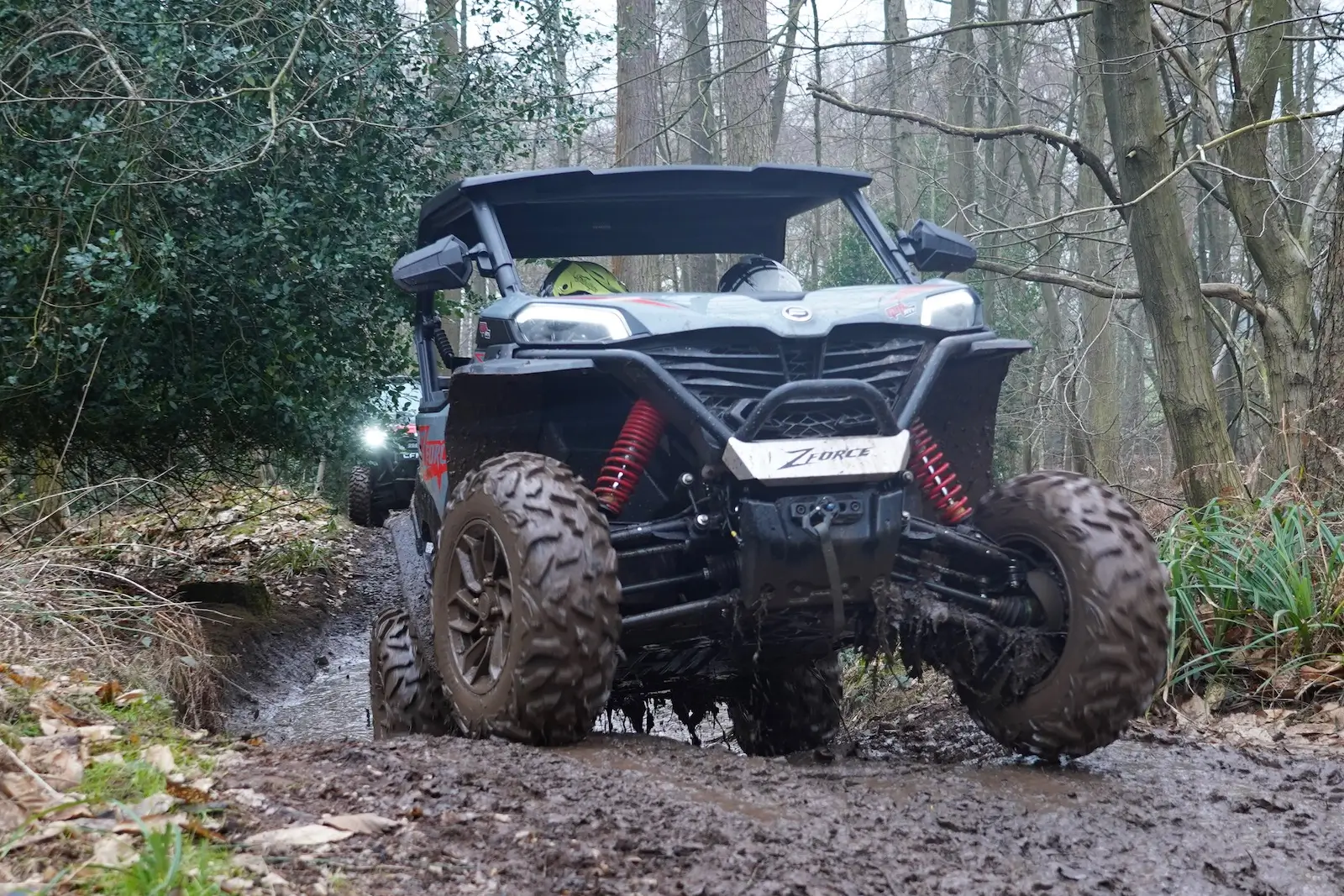A rugged off-road vehicle navigates a muddy forest trail with ease, its noticeable suspension and large tires reminiscent of a farm quad, cutting through the dense trees and lush foliage.