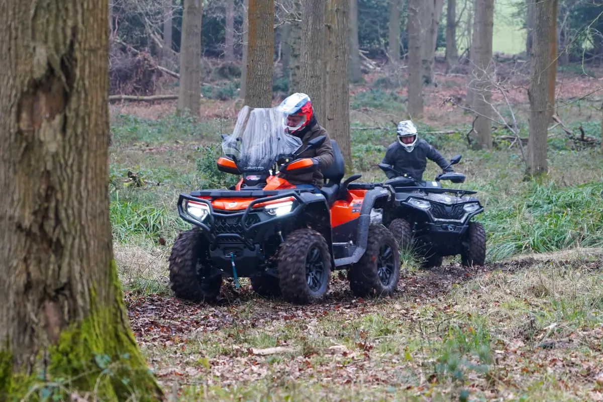 Two people ride CForce 850 Touring ATVs along a trail in a wooded forest area, surrounded by trees and lush vegetation.