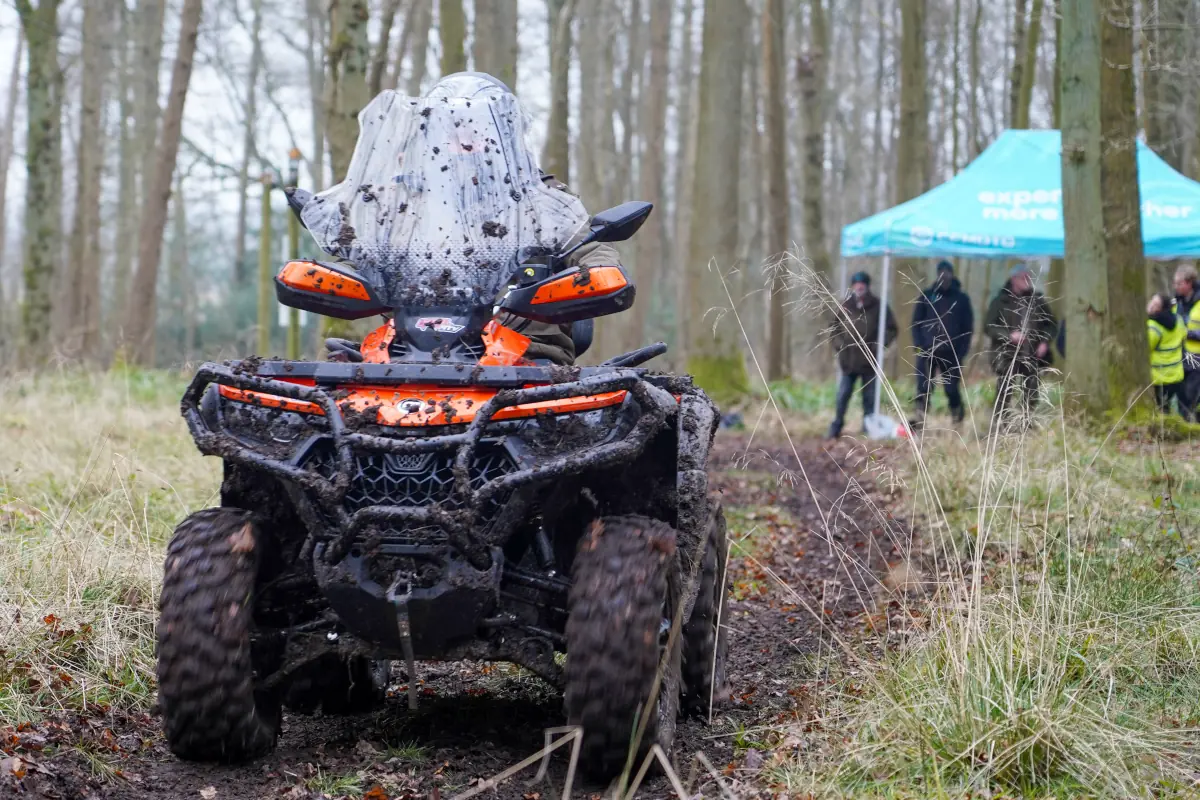 A person rides an orange and black CForce 850 Touring all-terrain vehicle through a muddy forest path, with mud splattered on the vehicle and rider; people and a blue tent are visible in the background.