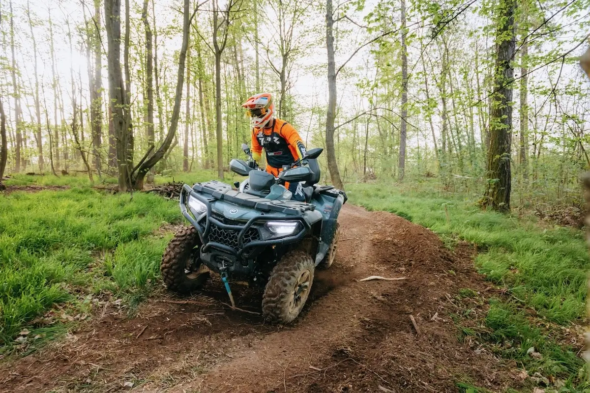 A person wearing an orange jacket and helmet rides a CForce 850 Touring ATV on a dirt trail through a forest with green grass and trees.