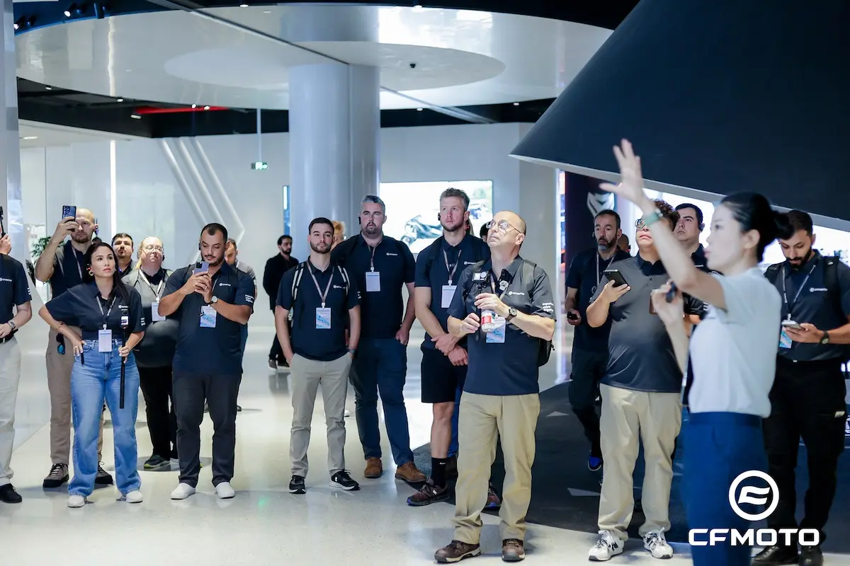 A group of people in matching shirts listen to a woman giving a presentation on ATV safety in a modern indoor space; the CFMOTO logo is visible in the corner.