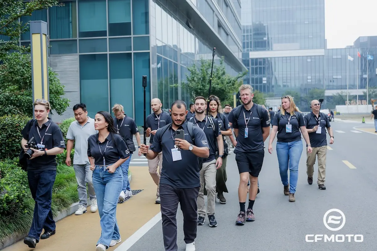 AHAI1161-opq4028661984 A group of people wearing matching CFMOTO shirts, promoting ATV safety, walk together outdoors near modern office buildings, some holding cameras and lanyards.