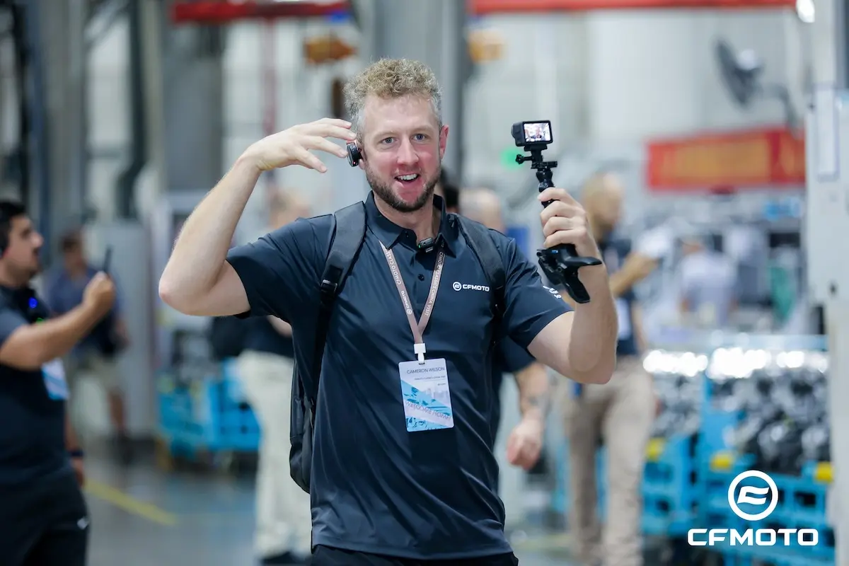 AHAI1181-opq4028648201 A man in a black CFMOTO shirt holds a small camera mounted on a handheld tripod while gesturing and smiling inside an industrial facility, possibly filming tips about ATV safety.