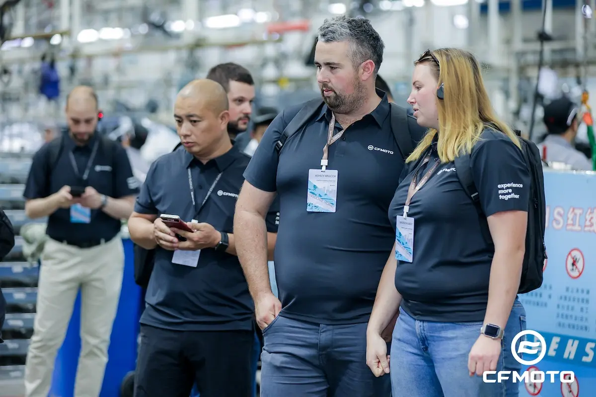 A group of people wearing matching CFMOTO shirts and name badges stand together indoors, some with headsets, in a factory or industrial setting, emphasizing the importance of ATV safety.