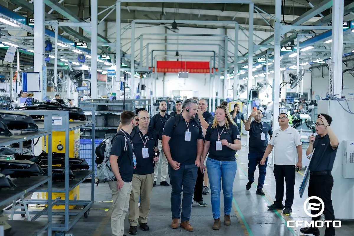A group of people wearing visitor badges tour a large, well-lit factory floor lined with machinery and equipment. The importance of ATV safety is highlighted as the CFMOTO logo appears in the lower right corner.