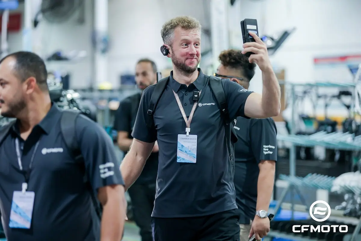 A man wearing a CFMOTO shirt and name badge takes a selfie with a smartphone in an indoor industrial setting, surrounded by others in similar attire, all promoting the importance of ATV safety.