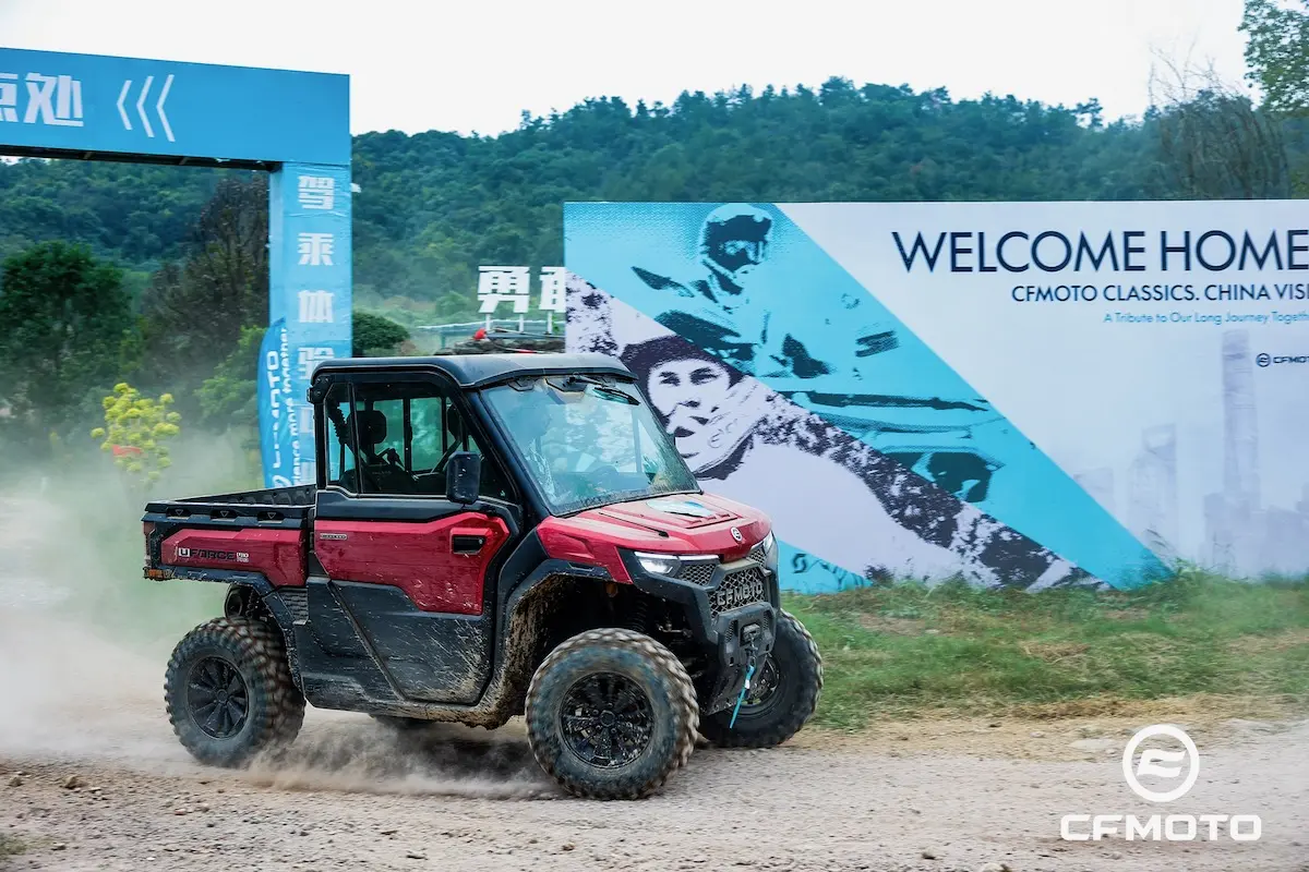 A red utility terrain vehicle drives on a dirt road near a large "Welcome Home" sign at an outdoor CFMOTO event, where ATV safety is always a top priority.