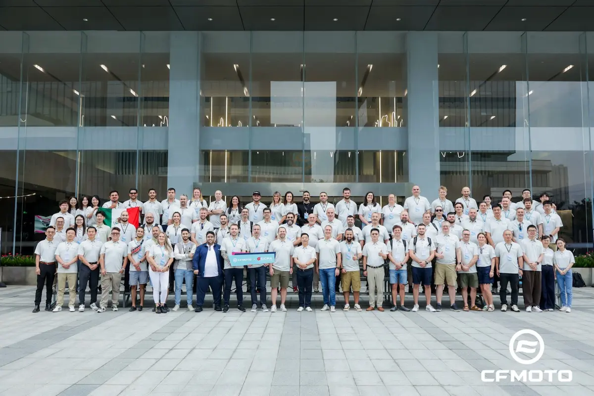 A large group of people stands in rows outside a modern building, posing for a group photo. Most are wearing light-colored shirts, reflecting their commitment to ATV safety. The CFMOTO logo appears in the corner.