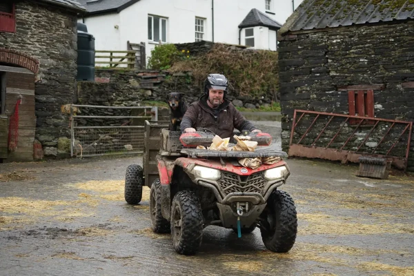 A man drives a quad bike loaded with firewood on a farm, with a dog sitting behind him. Stone buildings and a gated area appear in the background, raising practical questions like: what are OTR charges for such utility vehicles?.