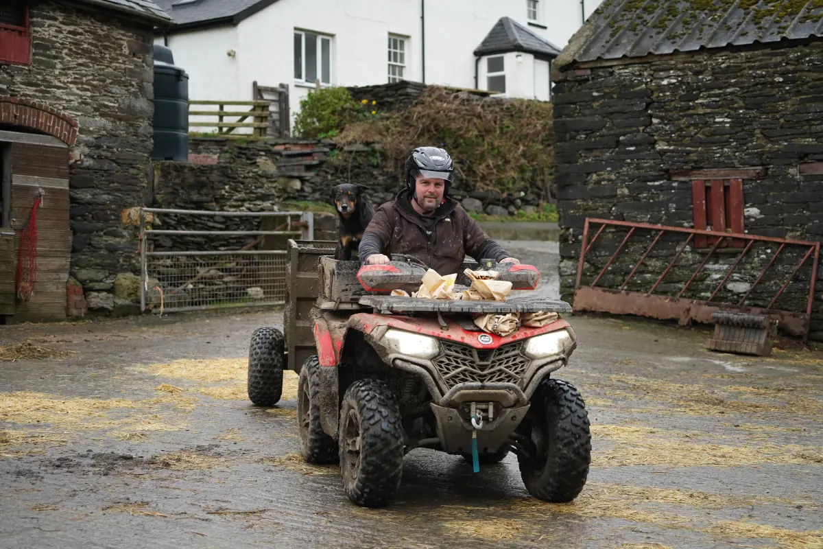 A man drives a quad bike loaded with firewood on a farm, with a dog sitting behind him. Stone buildings and a gated area appear in the background, raising practical questions like: what are OTR charges for such utility vehicles?.