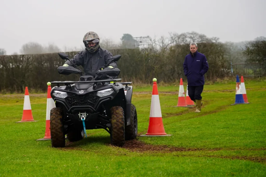 ATV safety training A person practices ATV safety by riding an ATV through a line of orange traffic cones on grass while another person stands nearby observing.