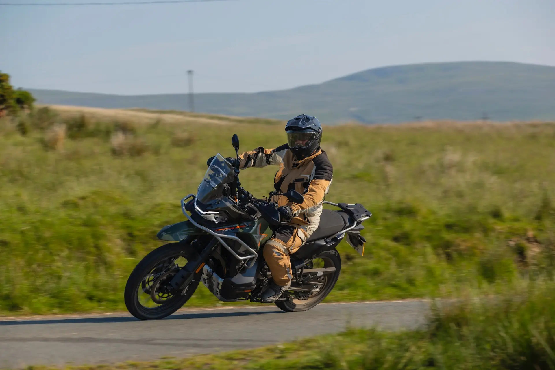 A person wearing tan and black motorcycle gear rides a black 1000MT-X adventure motorbike on a rural road with grassy hills in the background.