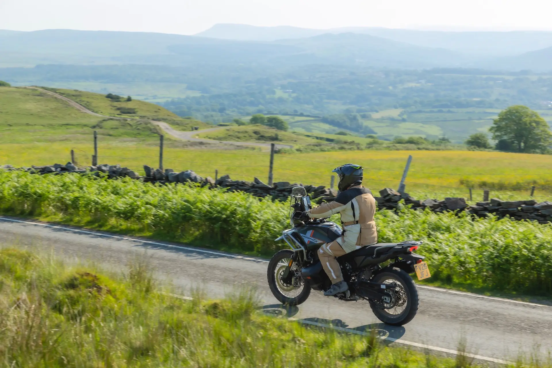 A person wearing protective gear rides a 1000MT-X motorbike down a country road, with fields, hills, and stone fences in the background.