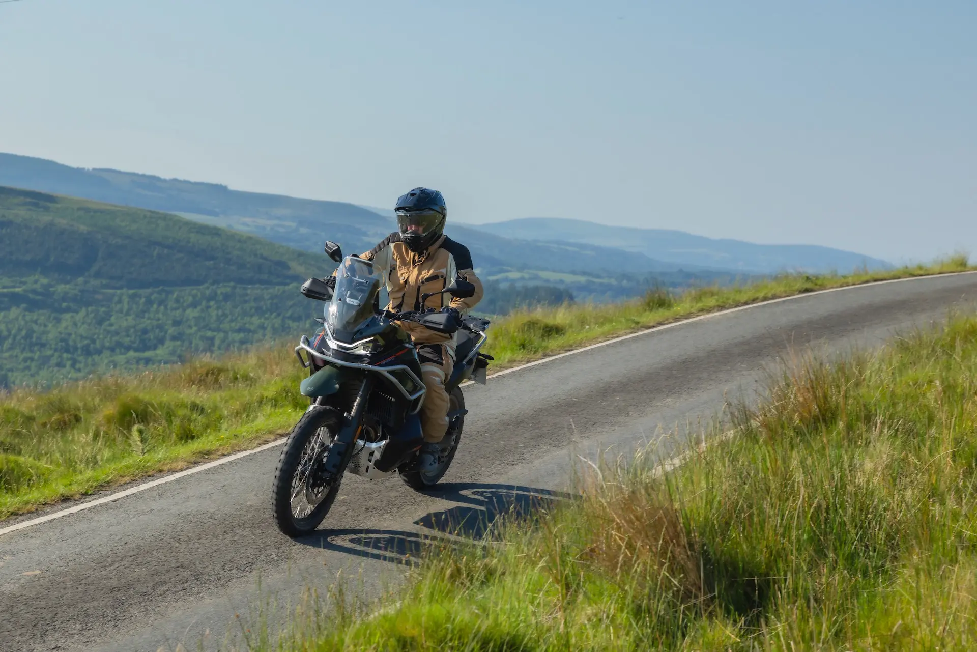 A person wearing protective gear rides a 1000MT-X motorbike on a narrow, winding road through a grassy, hilly landscape under a clear sky.