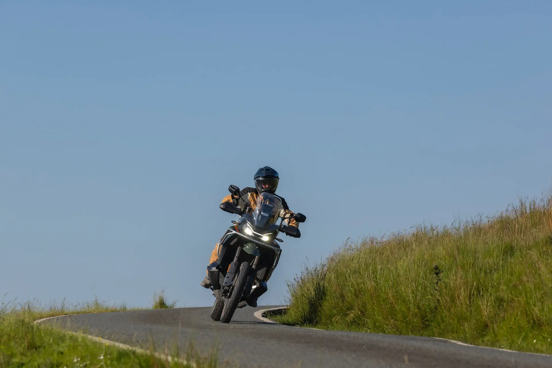 A person wearing a helmet rides a 1000MT-X motorbike on a paved road through green grassy hills under a clear blue sky.