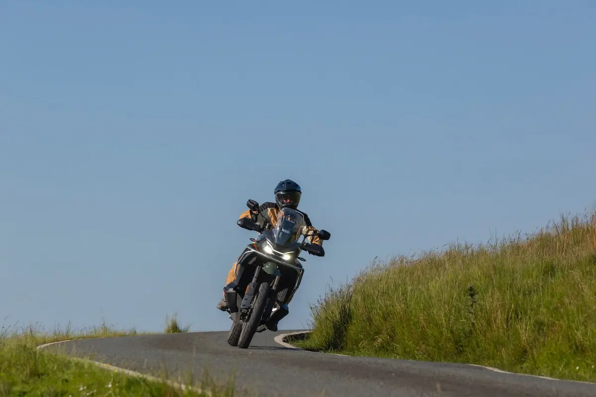 A person wearing protective gear rides a CFMOTO 1000MT-X on a curved, narrow road bordered by grass under a clear blue sky, capturing the spirit of adventure seen in any great CFMOTO 1000MT-X review.