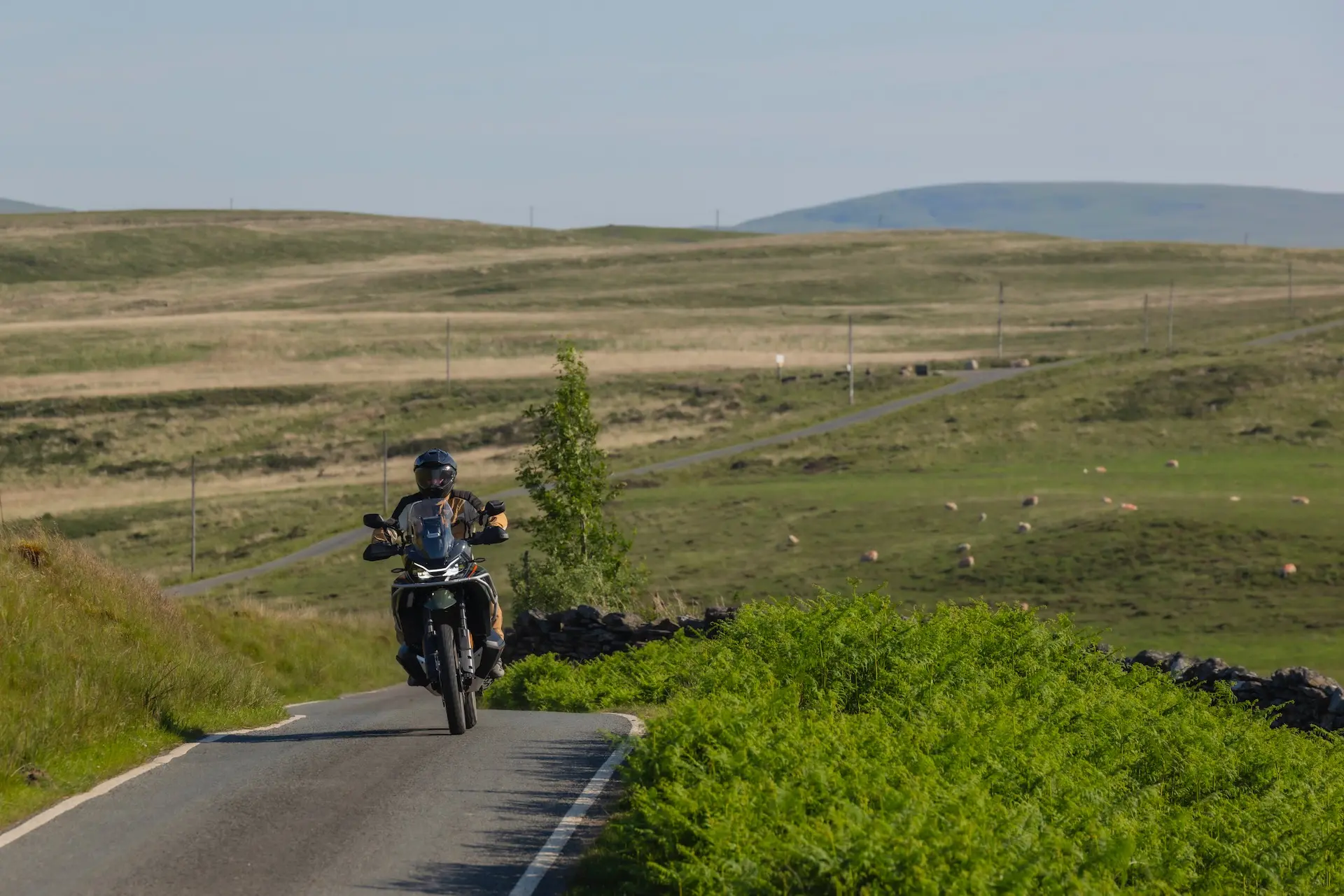 A person wearing a helmet rides a 1000MT-X motorbike on a narrow rural road, surrounded by green fields and distant hills under a clear sky.
