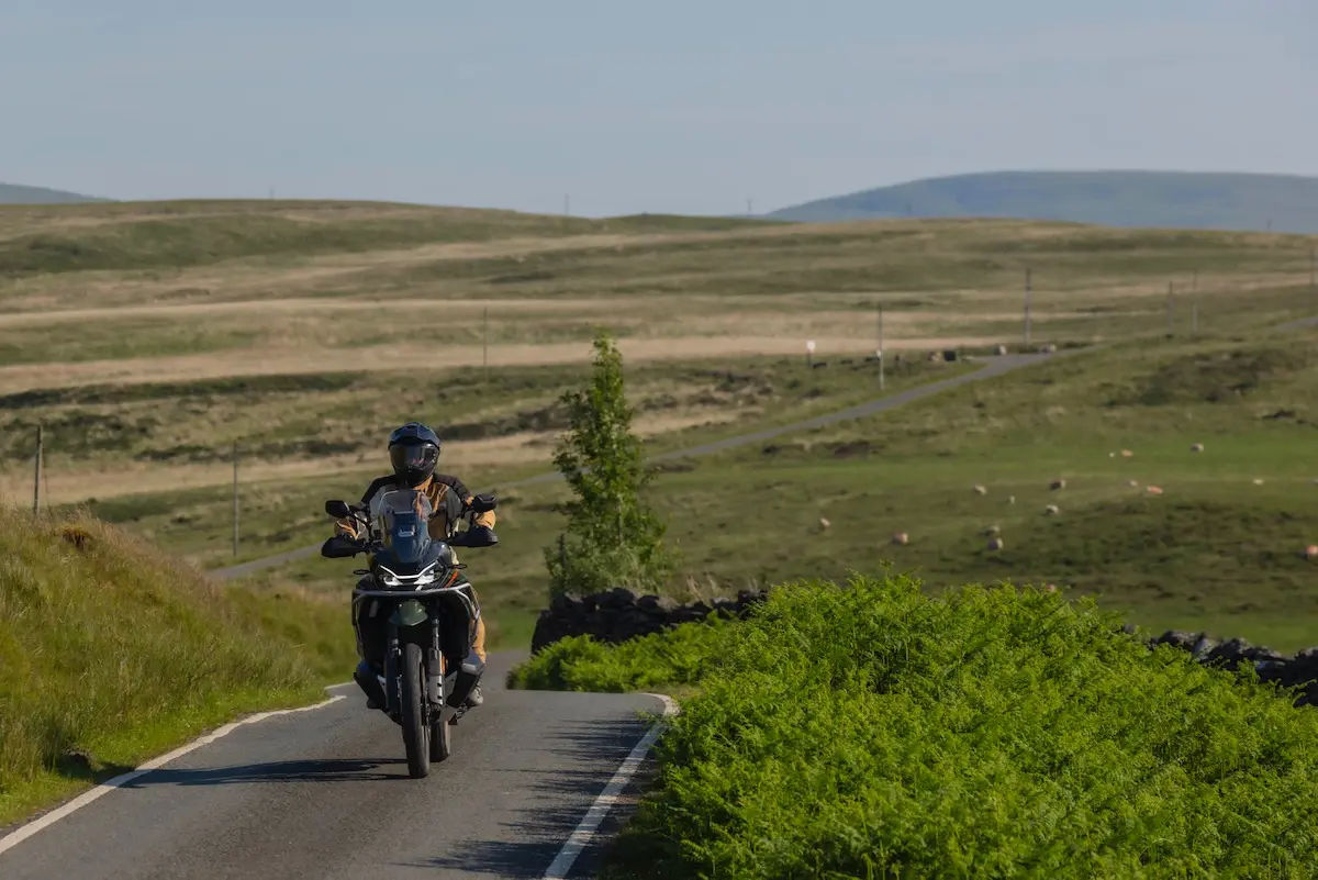 A person wearing a helmet rides a CFMOTO 1000MT-X motorbike on a narrow country road surrounded by grassy fields and distant hills under a clear sky, capturing the spirit of adventure found in any CFMOTO 1000MT-X review.