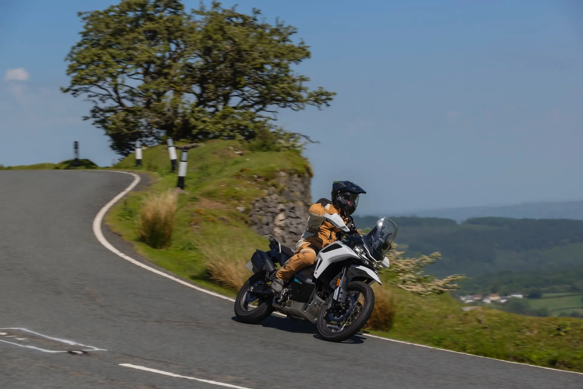 A person in tan clothing and a black helmet rides a 1000MT-X white motorbike around a bend on a rural road, framed by a tree and grassy landscape in the background.