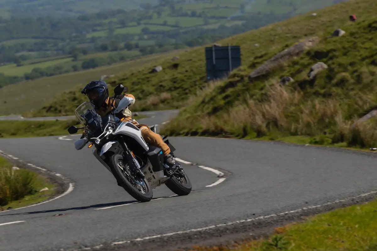 A person rides a CFMOTO 1000MT-X around a bend on a rural road, surrounded by green hills and an open landscape—capturing the spirit of adventure from any CFMOTO 1000MT-X review.