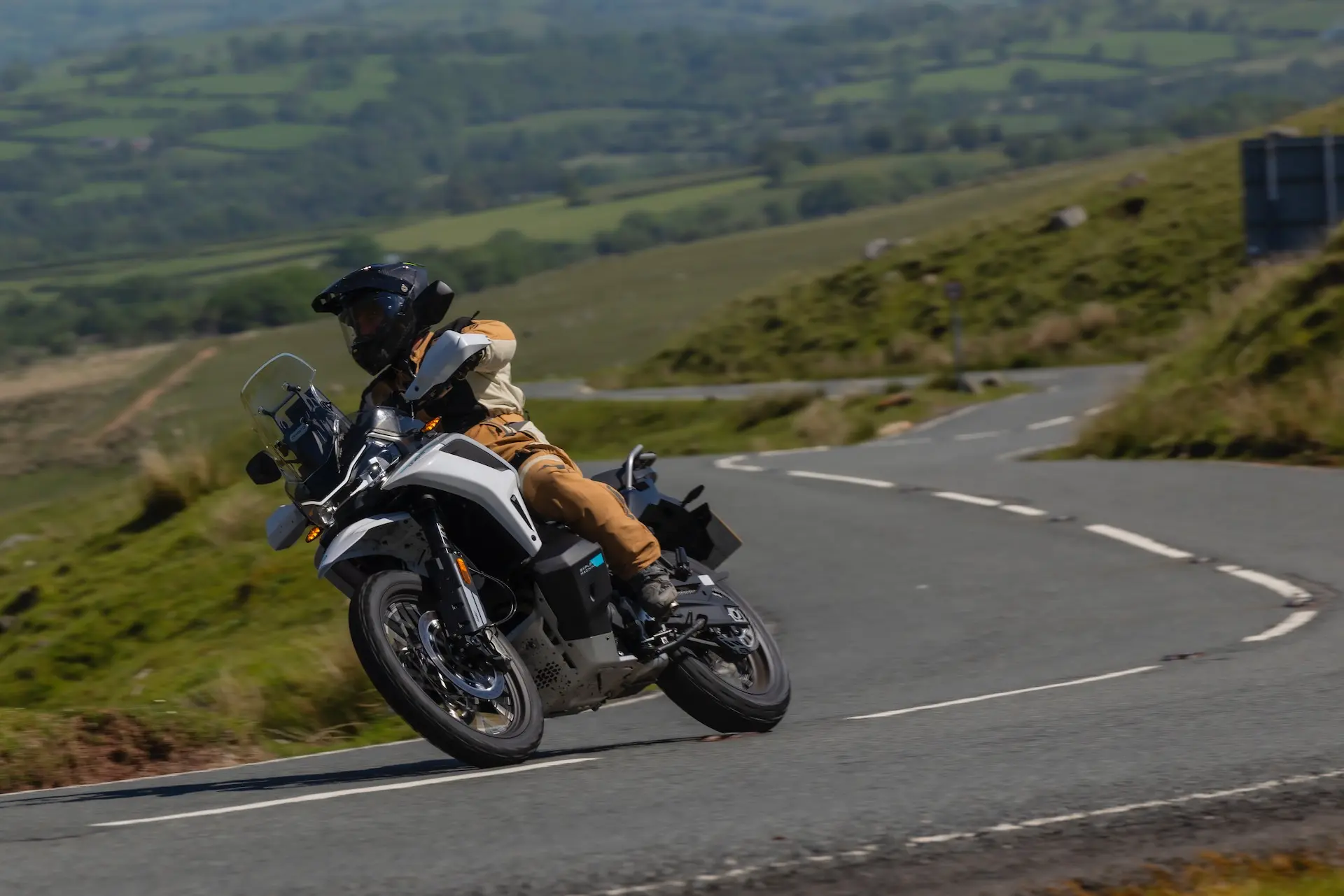 A person wearing protective gear rides a 1000MT-X white motorbike round a bend on a rural road with green hills in the background.