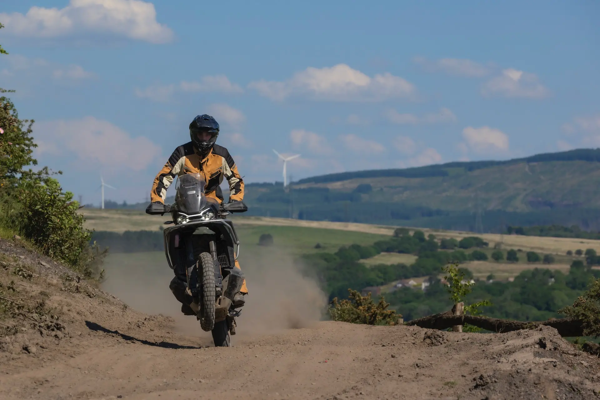 A person in protective gear rides a 1000MT-X motorbike on a dusty dirt track through a rural landscape with hills and wind turbines in the background.