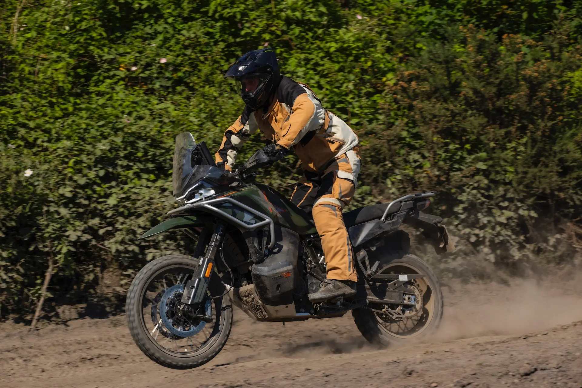 A person in tan and black riding gear rides a 1000MT-X adventure motorbike on a dirt track, surrounded by greenery and dust.