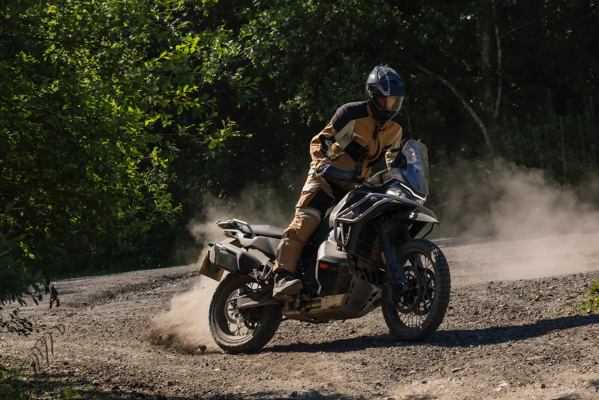 A person wearing protective gear rides a 1000MT-X motorbike on a gravel road, kicking up dust whilst turning, with trees in the background.