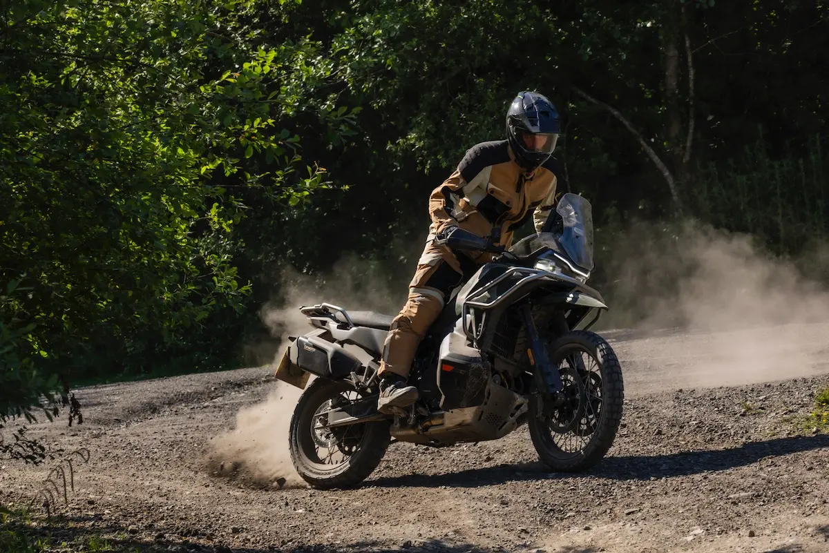 A person wearing protective gear rides a CFMOTO 1000MT-X motorbike on a gravel road, kicking up dust whilst making a turn near a wooded area—a scene that could headline any CFMOTO 1000MT-X review.