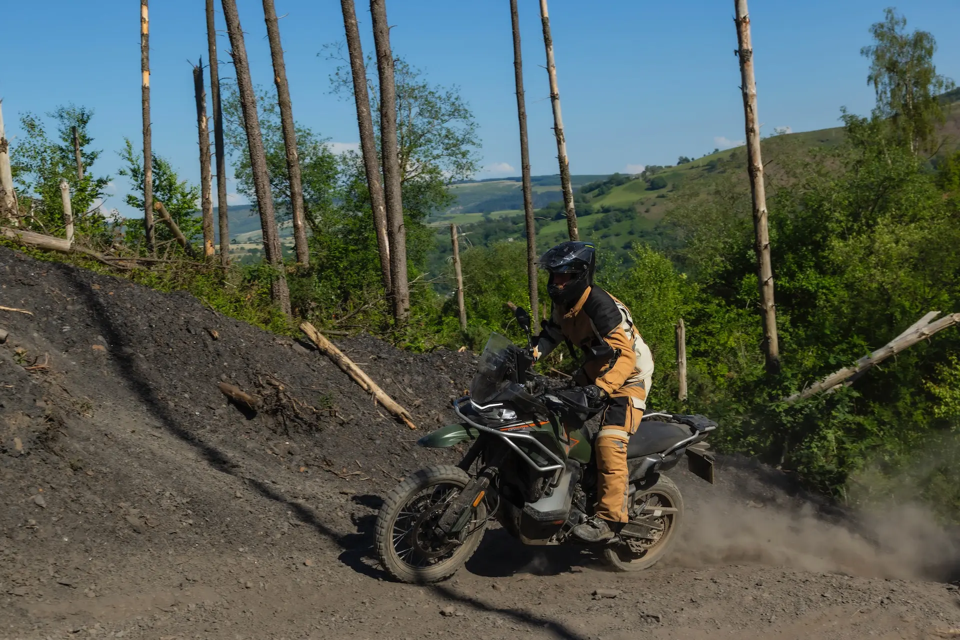 A person in protective gear rides a 1000MT-X motorbike up a dirt track in a hilly, forested area on a sunny day.