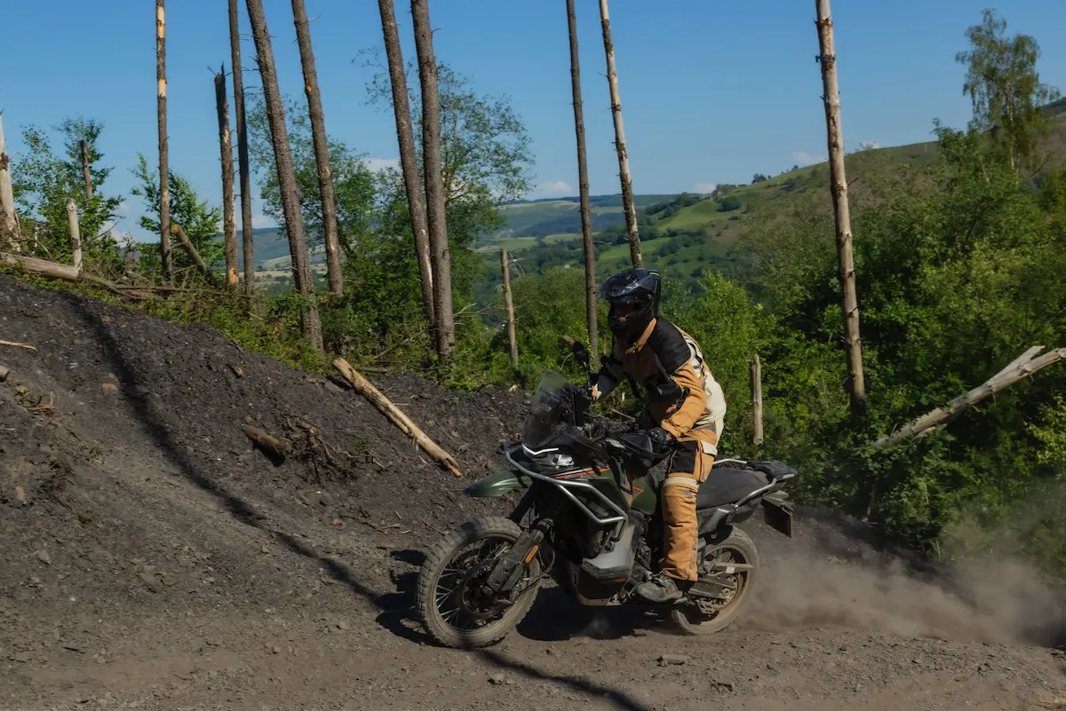 A person wearing protective gear rides a CFMOTO 1000MT-X motorcycle on an off-road trail through a wooded, hilly landscape on a sunny day, capturing the spirit of adventure in this CFMOTO 1000MT-X review.