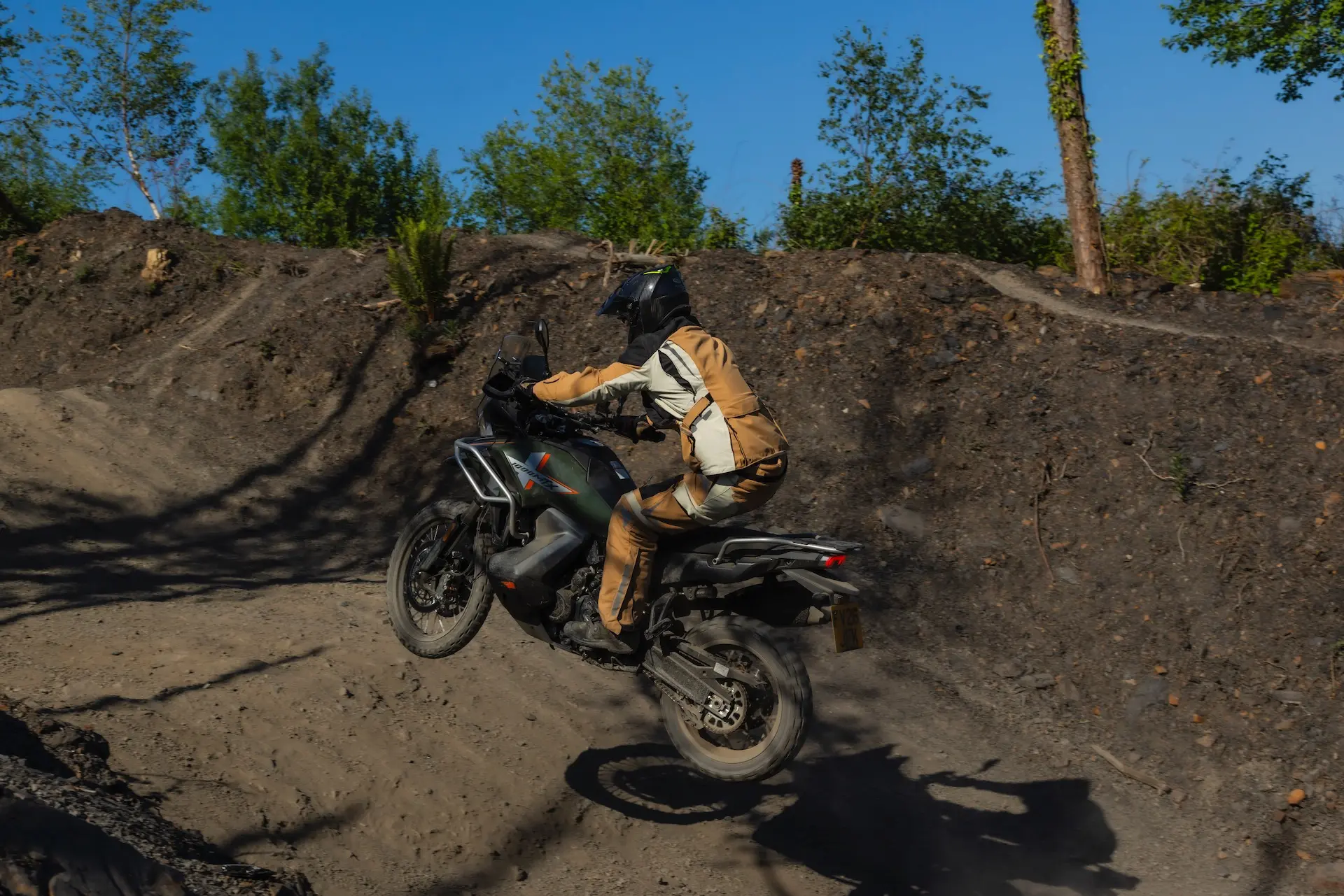 A person in protective gear rides a 1000MT-X motorbike off-road, kicking up dust on a dirt track surrounded by trees and shrubs under a clear blue sky.