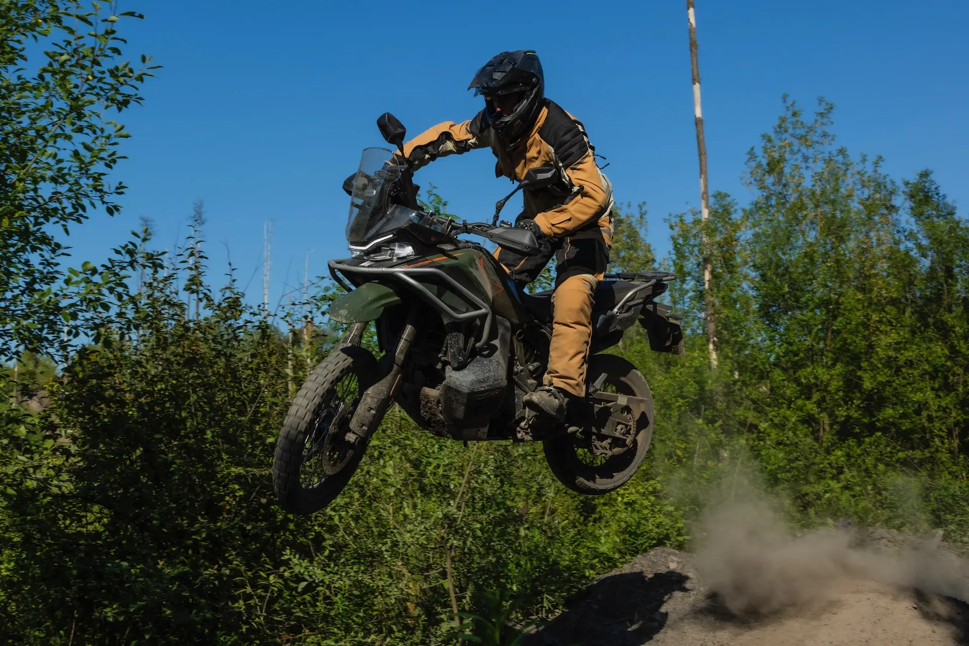 A person in protective gear rides a 1000MT-X motorbike off-road, catching air over a dirt mound with dust trailing behind. Trees and greenery are visible in the background.