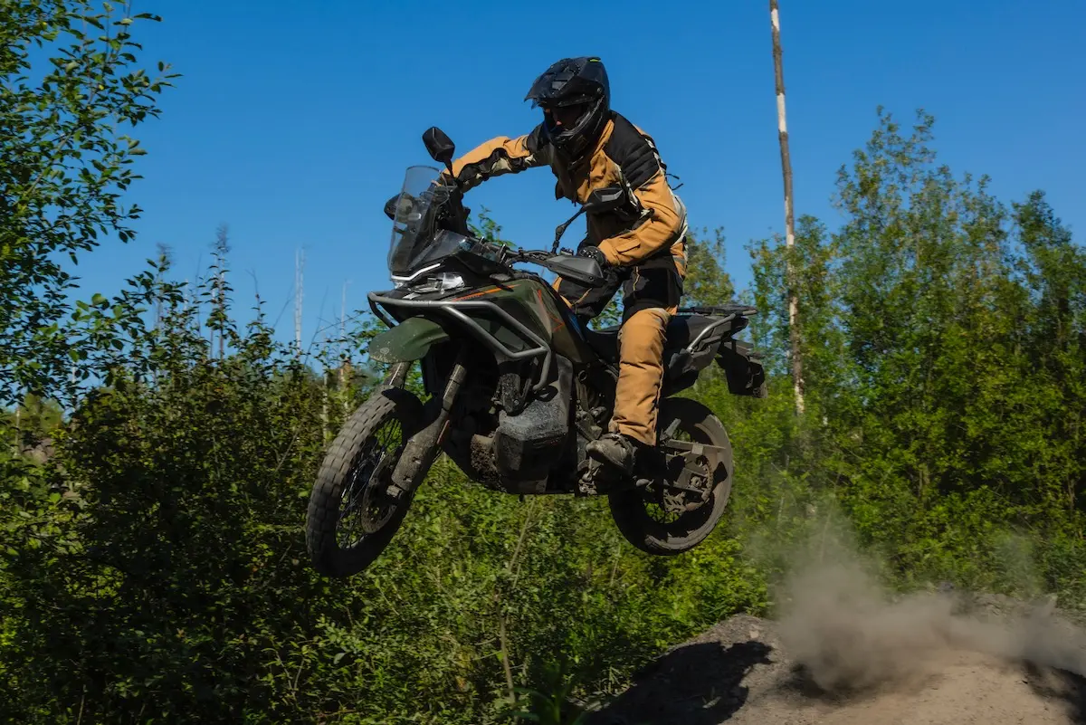 A person in protective gear rides a CFMOTO 1000MT-X off-road, airborne above a dirt track with trees and greenery in the background—perfect for an action-packed CFMOTO 1000MT-X review.