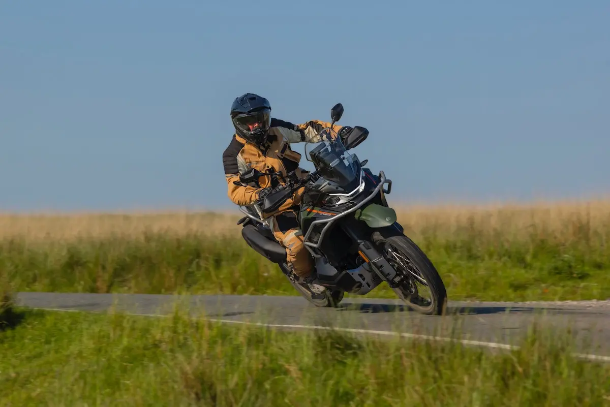 A motorcyclist in protective gear leans into a bend on a tarmacked road, riding the CFMOTO 1000MT-X through a grassy landscape under a clear sky—a scene straight out of a CFMOTO 1000MT-X review.