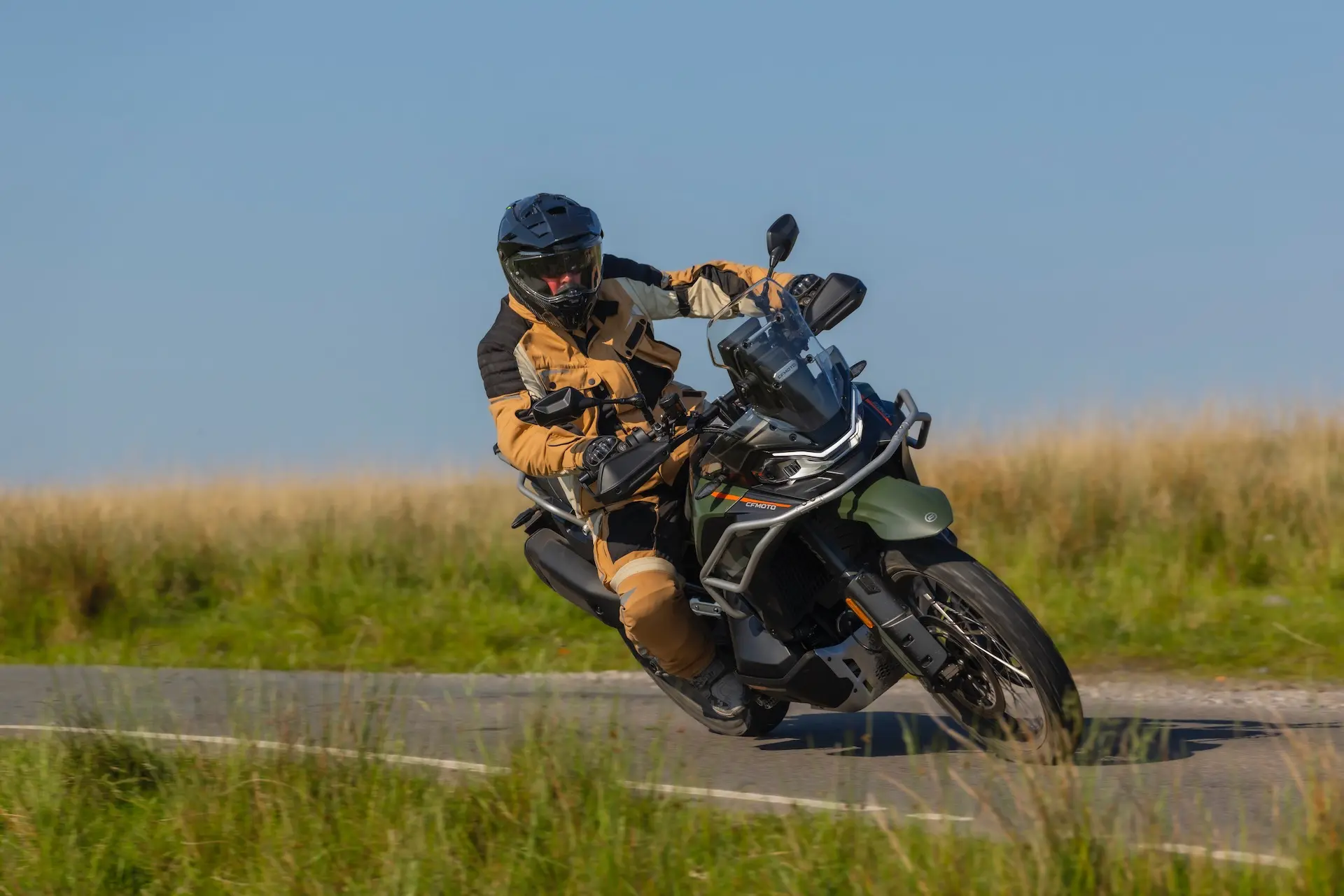 A person in protective gear rides a green 1000MT-X adventure motorbike around a bend on a rural road with grassy fields in the background.