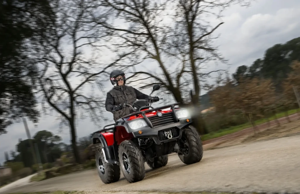 A person is riding a red farm quad along a rural path, surrounded by bare trees in the background, wearing a helmet and dark clothing.