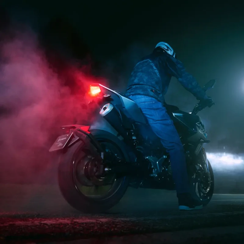 A person wearing a helmet sits on a CFMoto motorcycle at night, with red taillights and exhaust smoke illuminated by surrounding lights.