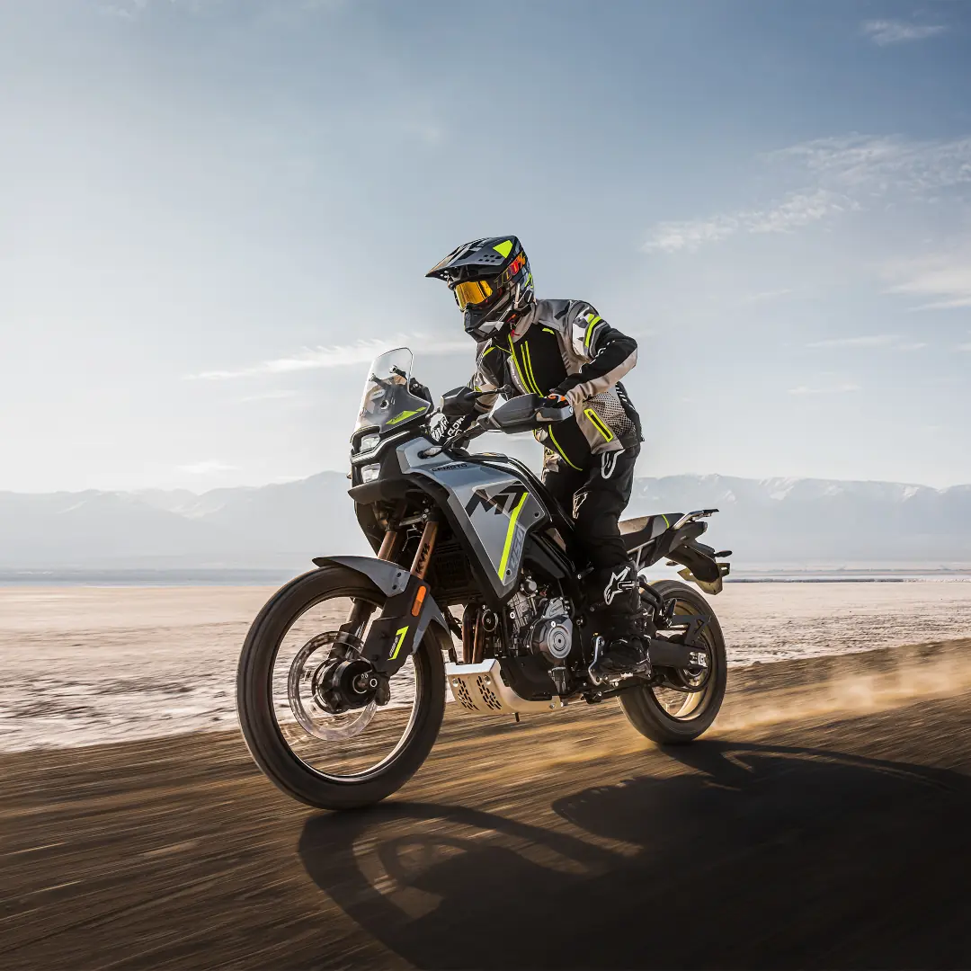 A person wearing full riding gear rides a black and yellow 450MT adventure motorcycle on a dirt road with mountains in the background.