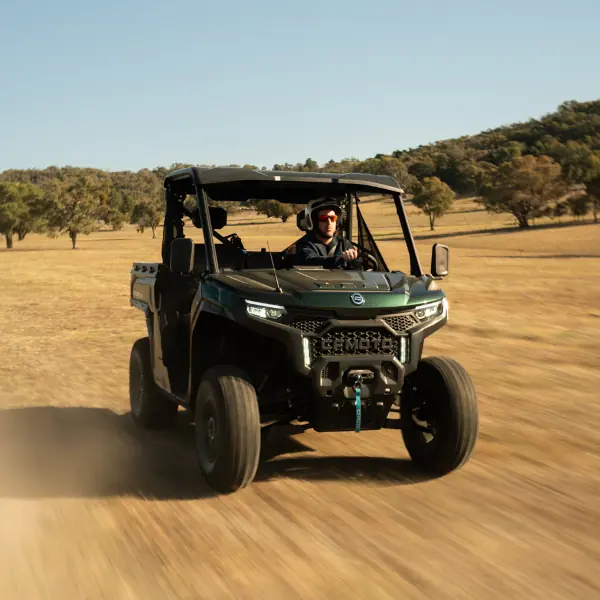 A person drives a dark green CFMOTO Agri Range utility terrain vehicle (UTV) across a dry, grassy field with trees and hills in the background under a clear sky.