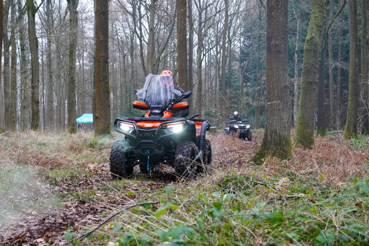 Two people ride CForce 850 Touring all-terrain vehicles along a dirt path through a forest with bare trees and scattered foliage on the ground.
