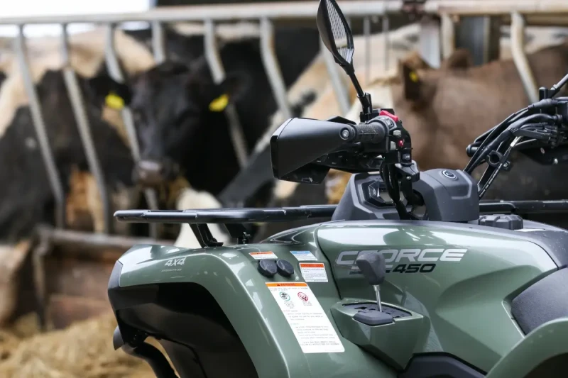 CFMOTO CFORCE ATV A close-up of a green CFORCE 450 ATV equipped with a CFMOTO T-Box, parked near a group of cows behind a metal fence.