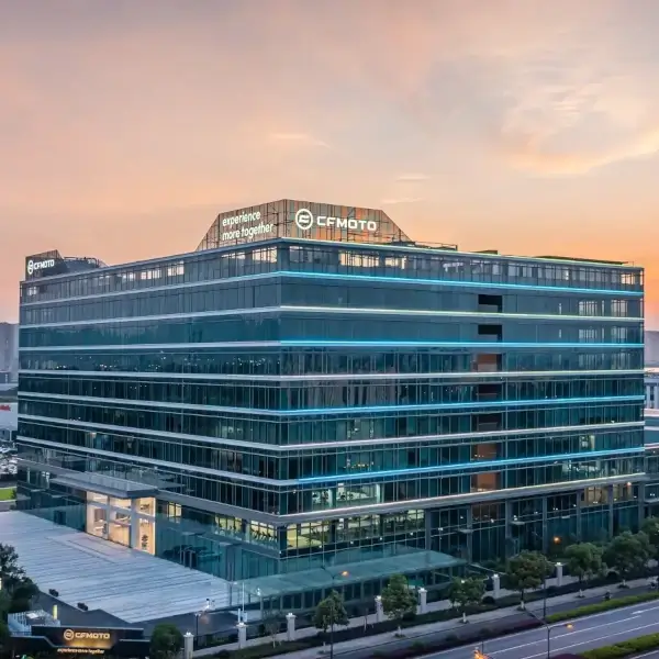 A modern glass office building with “CFMOTO” signage on the roof, photographed at sunset with some trees and a road in the foreground.