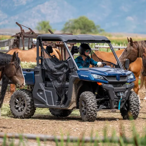 A person wearing a helmet drives a blue side-by-side utility vehicle near several equestrian horses in a fenced outdoor area.