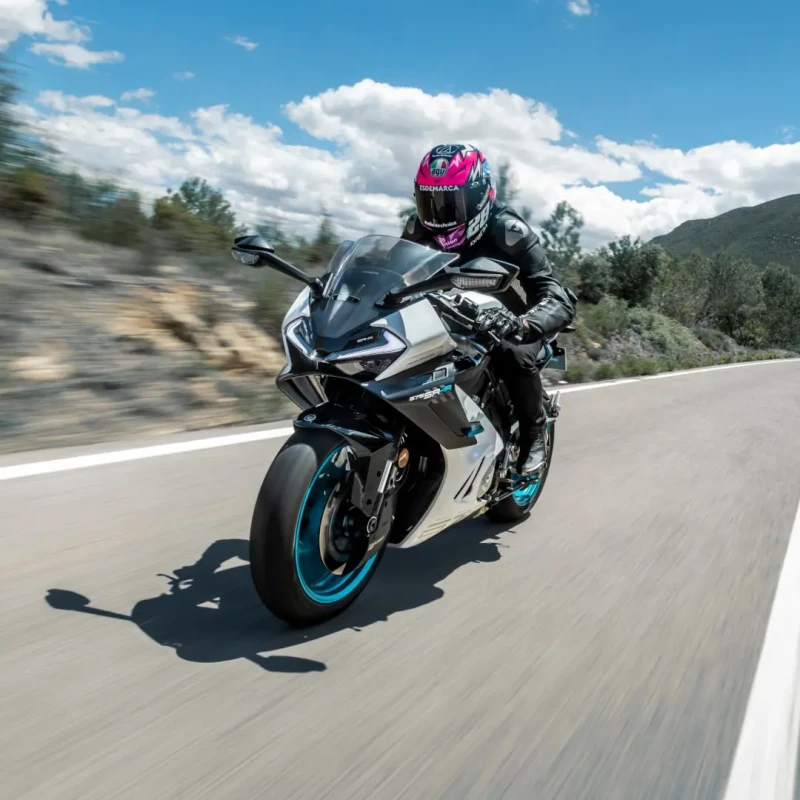 A motorcyclist in black gear and a pink helmet rides a silver CFMoto motorcycle at speed on a paved road, with trees and hills in the background.