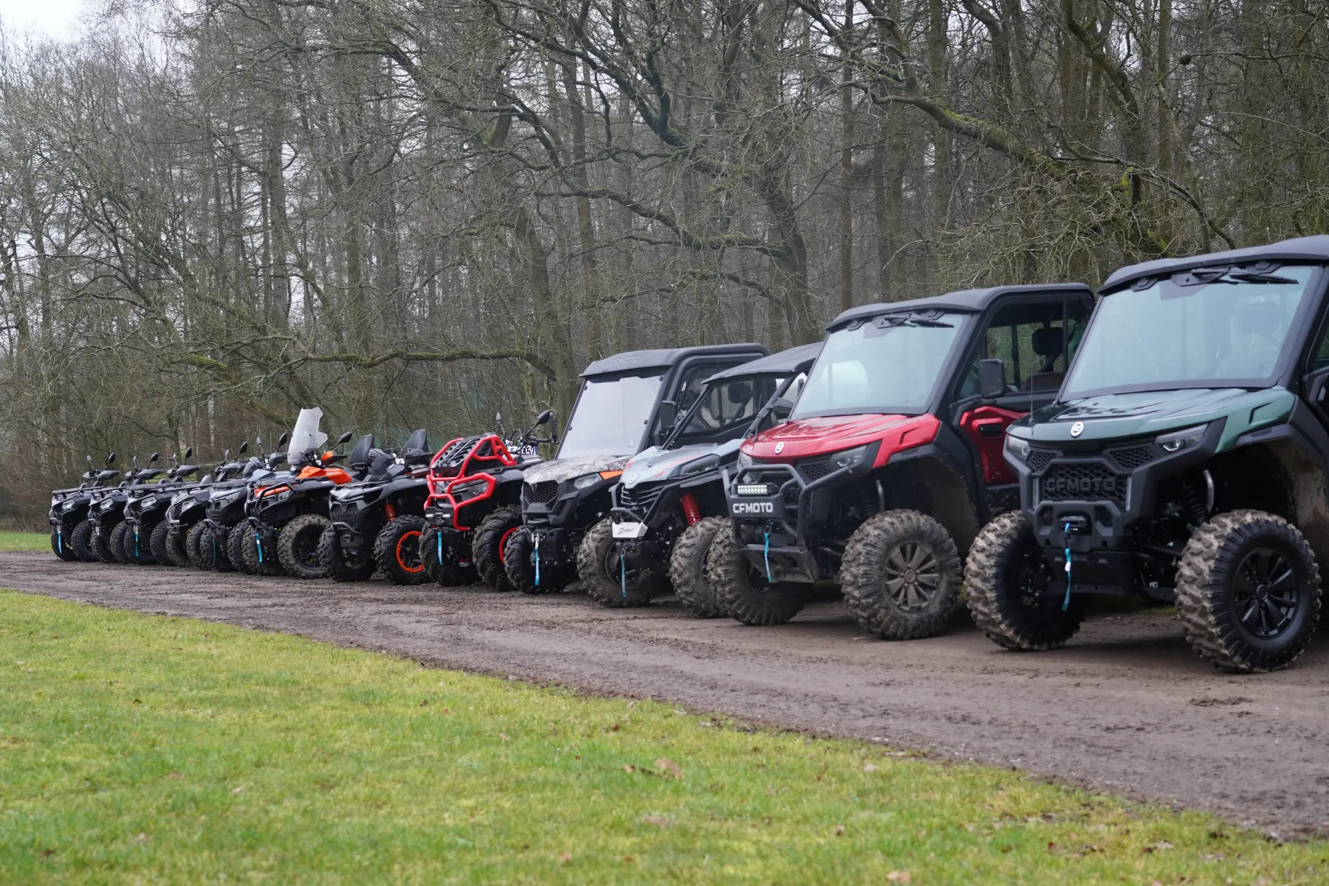 A row of off-road vehicles, including ATVs, UTVs, and CFMOTO models, parked on a dirt path beside a grassy area with leafless trees in the background.